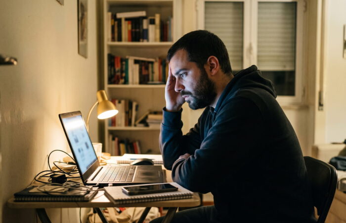 Homem sentado a uma secretária num apartamento em Portugal, à noite, a olhar para o ecrã de um portátil com expressão cansada, telemóvel ao lado