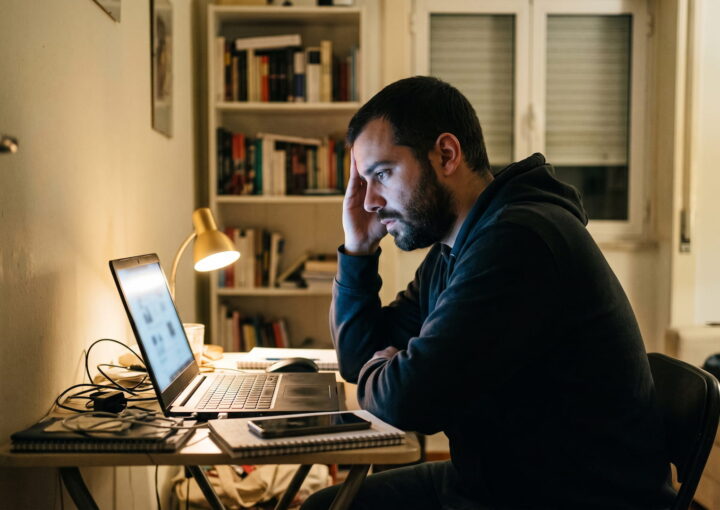 Homem sentado a uma secretária num apartamento em Portugal, à noite, a olhar para o ecrã de um portátil com expressão cansada, telemóvel ao lado