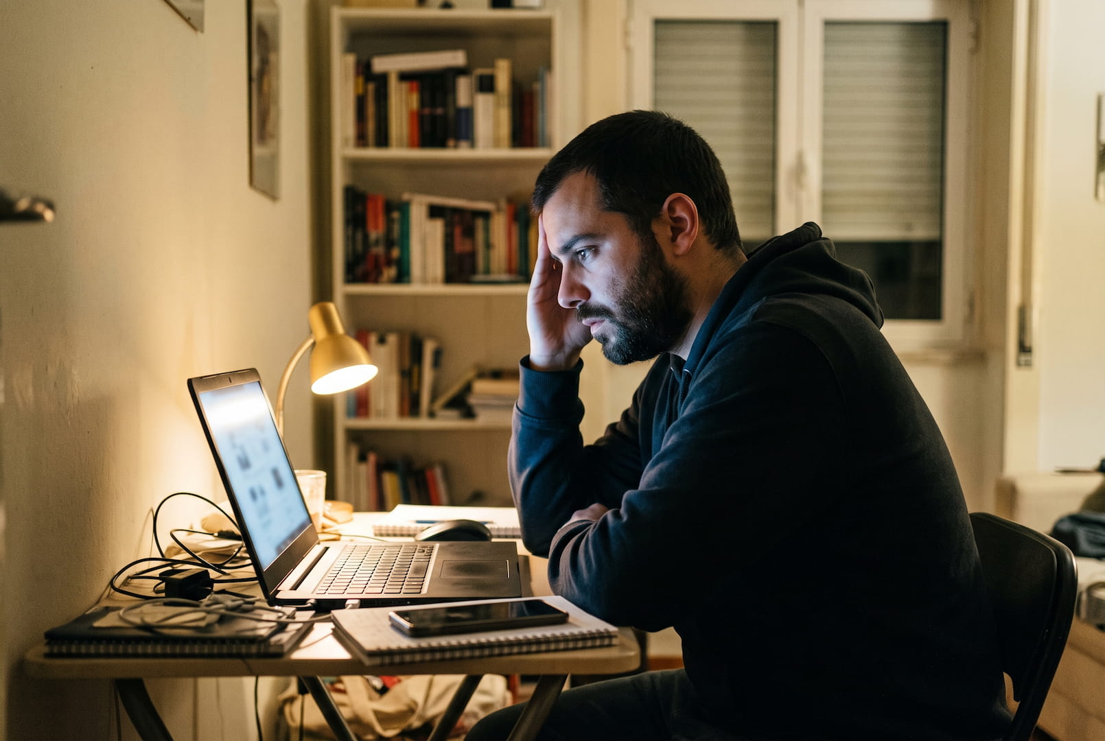 Homem sentado a uma secretária num apartamento em Portugal, à noite, a olhar para o ecrã de um portátil com expressão cansada, telemóvel ao lado