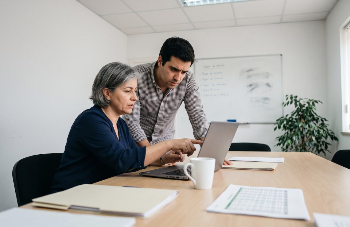 Duas pessoas numa pequena sala de reuniões de uma empresa, uma delas aponta para um portátil